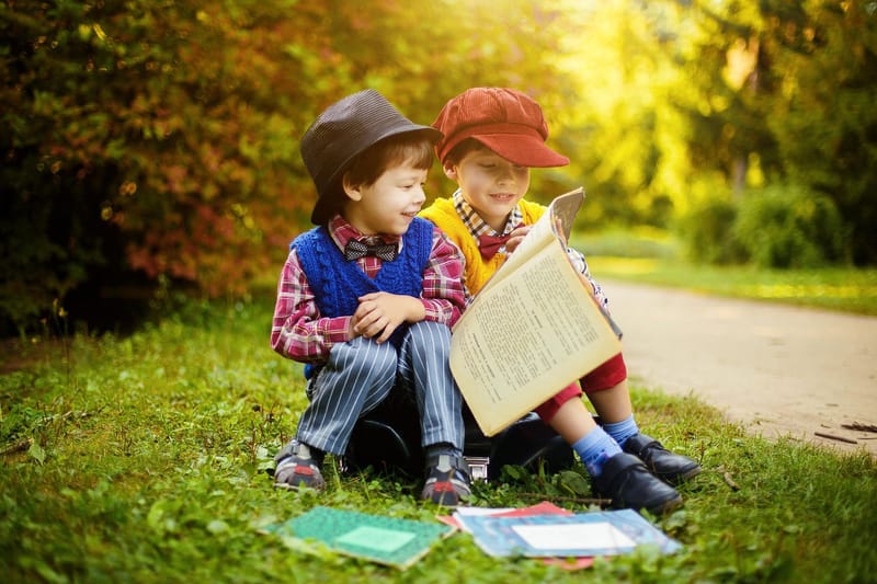 Two young boys sitting together on the grass, smiling as they read the same book
